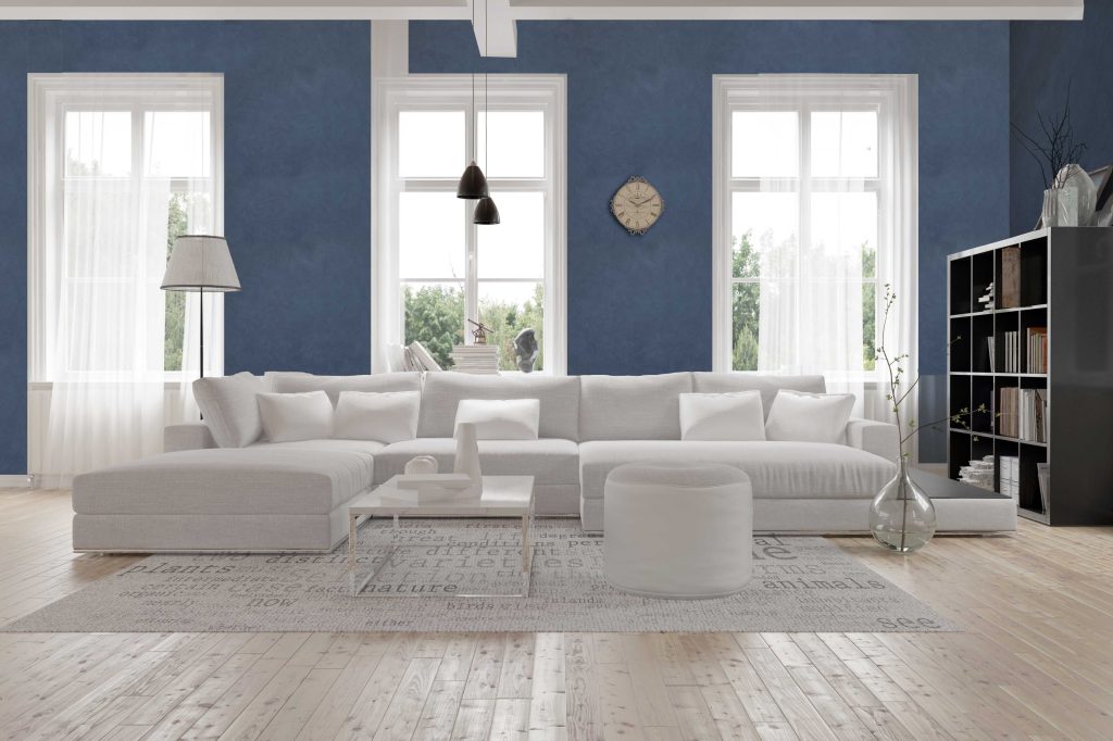 Modern spacious lounge or living room interior with monochromatic white furniture and decor below three tall bright windows with a dark bookcase accent in the corner